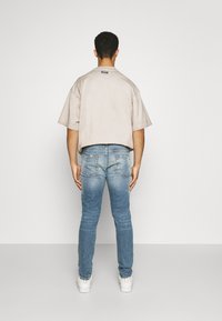 Camiseta beige oversized de manga corta, combinada con jeans skinny azules desgastados. Zapatillas blancas completan el atuendo. Vista trasera del portador.