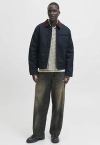 Marineblauer Bomberjacke mit einem braunen Cordkragen, darunter ein kariertes Hemd und ausgebleichte Schlagjeans. Das Model trägt dunkles Schuhwerk.