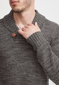 Grau gestrickter Pullover mit großem Schalkragen. Verfügt über einen Holzknopf am Kragen und gerippte Bündchen. Die Textur ist weich und dick.