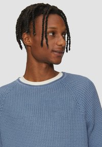 Pull en maille bleu clair avec un motif textur�é, col rond et manches raglan, superposé sur un sous-vêtement blanc.