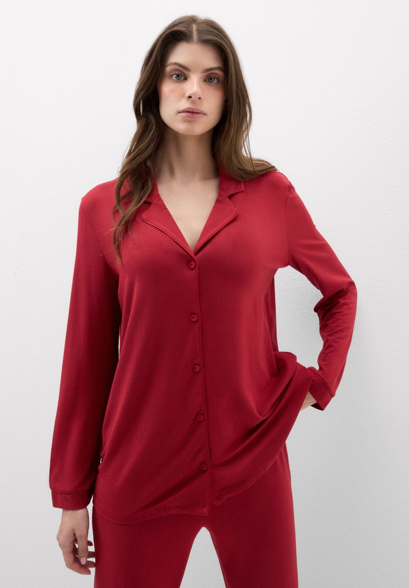 Mujer con cabello largo y castaño viste un top rojo de manga larga con botones y pantalones a juego, de pie con una mano en el bolsillo.