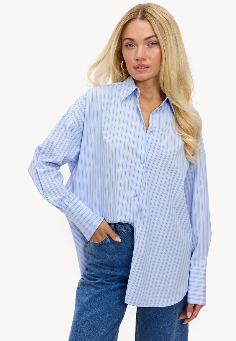 Femme blonde portant une chemise boutonnée à rayures bleu clair et un jean bleu, posant avec une main dans la poche sur un fond blanc.