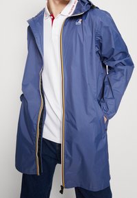 Blå vandtæt jakke med hætte, der har en lynlås foran med gul accent, letvægtsmateriale og sidelommer.