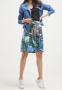 Une veste en denim sur un haut noir, associée à une jupe longueur genou avec un motif tropical bleu et vert. Des baskets blanches complètent le look.