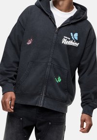 Felpa nera con zip e cappuccio regolabile. Presenta un ricamo di farfalla colorato in rosa e verde. Materiale in cotone, tasche frontali.