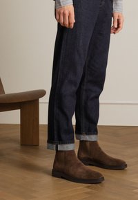 Stivali alla caviglia in suede marrone con punta arrotondata e suola piatta in gomma, indossati con jeans in denim scuri risvoltati. Mobili in legno e pavimenti sullo sfondo.