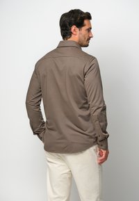 Camicia marrone a maniche lunghe con taglio dritto e polsini abbottonati, realizzata in tessuto liscio, abbinata a pantaloni color crema.