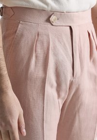 Pantalones de lino rosa claro con pliegues, cierre de botón y cintura ajustada. Presentan una textura suave y bolsillos laterales.