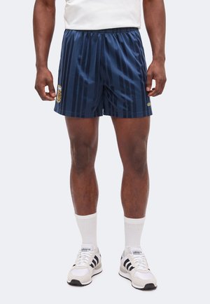 Bărbat care poartă pantaloni scurți sportivi cu dungi bleumarin, cu stema Argentinei și logo-ul Adidas, șosete albe și adidași albi Adidas.