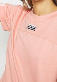 Ljust rosa bomullströja med rund halsringning, korta ärmar och en svart och vit Adidas-logotyp på bröstet.