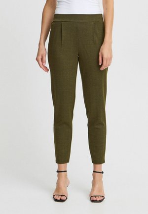 Femme portant un pantalon slim vert olive avec un motif chevrons subtil et des sandales à talons hauts noires à bride cheville, debout sur fond blanc.