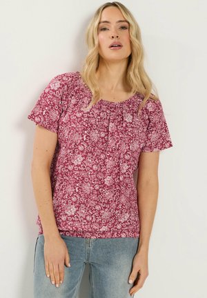 Pitkähiuksinen vaaleatukkainen nainen, jolla on punainen lyhythihainen kukkakuvioinen paita ja vaaleansiniset farkut, seisoo yksinkertaista valkoista seinää vasten.