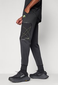 Sorte atletiske joggers med en side lomme med et sort afsnit og grønne geometriske linjer. Sættet er kombineret med sorte sko, hvilket fremhæver det strømlinede design.