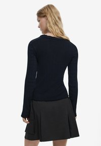 Sötétkék bordázott, hosszú ujjú felső, amely testhez simuló siluettel és kerek nyakkivágással rendelkezik, fekete, redőzött szoknyával párosítva, amely oldalzsebekkel van ellátva.