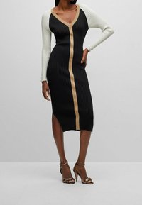 Robe tricotée avec une base noire, des manches blanches et une bande verticale dorée. Design en V avec texture côtelée et détail d'une fente sur le côté.