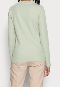 Top a maniche lunghe a coste di un delicato verde menta, realizzato in tessuto morbido con un design aderente e una texture sottile, abbinato a pantaloni beige.