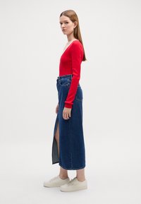 Rød langermet topp kombinert med et høyt liv denim maxiskjørt med splitt i siden, komplementert av beige sneakers. Glatte teksturer i stoffet synlige.