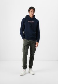 Marineblauwe hoodie met trekkoord, voorzien van een logo op de voorkant, gecombineerd met olijfgroene cargobroeken en witte sneakers. Eenvoudige, casual outfit.
