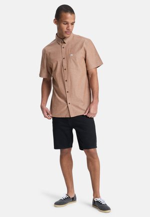 Koszula z krótkim rękawem w jasnobrązowym kolorze, zapinana na guziki, z kieszenią na piersi, ciemne szorty i szare buty z białymi sznurowadłami, stojące na jednolitym tle.