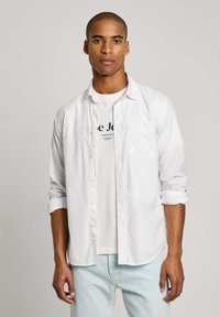 Camicia bianca a maniche lunghe con bottoni e tasca, indossata sopra una maglietta bianca con grafica, abbinata a jeans azzurri chiari. Texture del tessuto liscia.