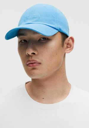 Jonge man met lichte huid en sproeten, draagt een lichtblauwe Lululemon-pet en een effen wit T-shirt met ronde hals, kijkt iets naar links.