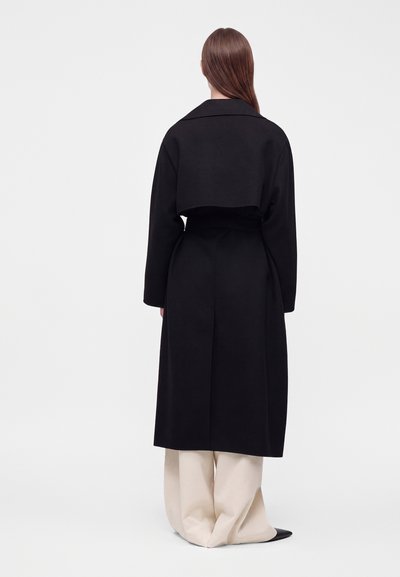 Femme aux longs cheveux bruns portant un manteau long noir sur un pantalon beige large et des chaussures noires pointues, debout devant un fond clair uni.