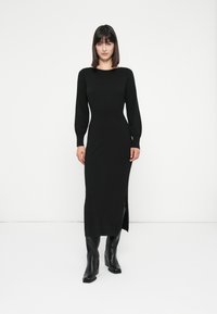 Donna in un abito midi nero a maniche lunghe aderente con spacco laterale, in piedi davanti a uno sfondo bianco, indossando stivali con tacco neri.