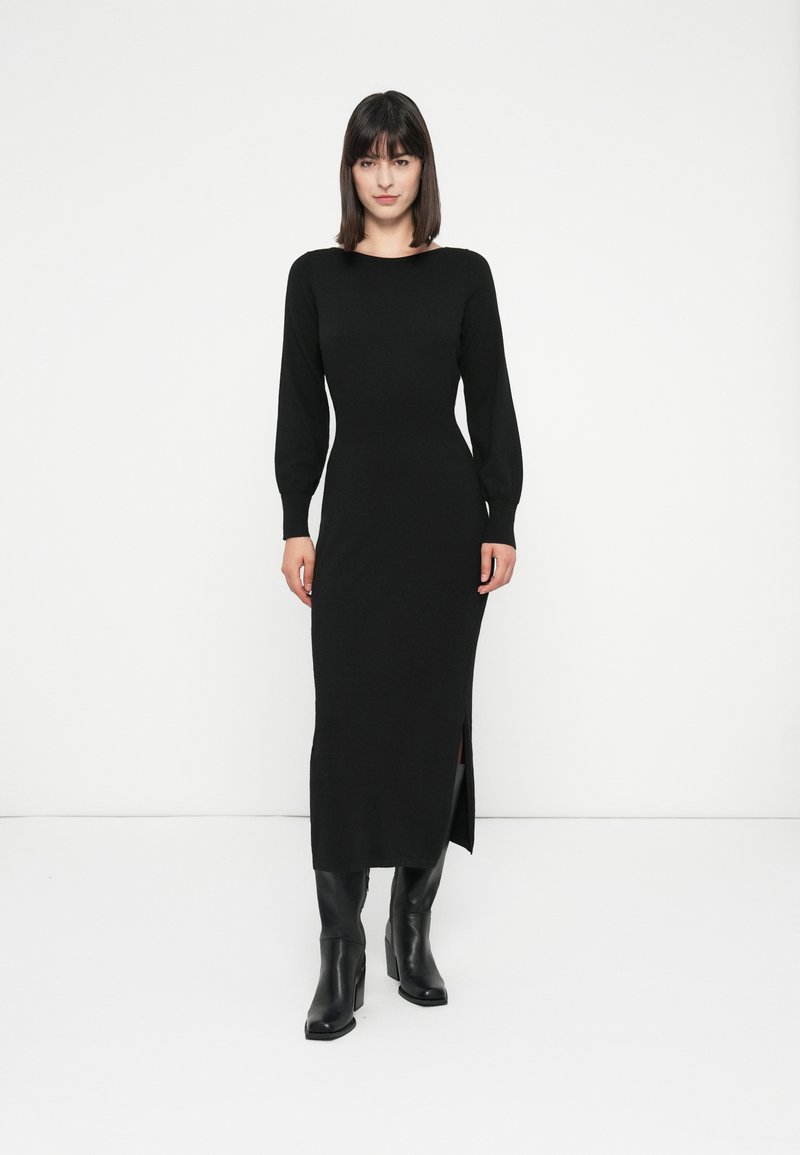 Donna in un abito midi nero a maniche lunghe aderente con spacco laterale, in piedi davanti a uno sfondo bianco, indossando stivali con tacco neri.