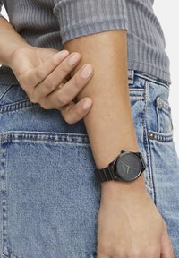 Schwarze analoge Uhr an einem Handgelenk, mit einem runden Ziffernblatt und einfachen Markierungen, kombiniert mit einem schwarzen Metallarmband. Denimstoff im Hintergrund.