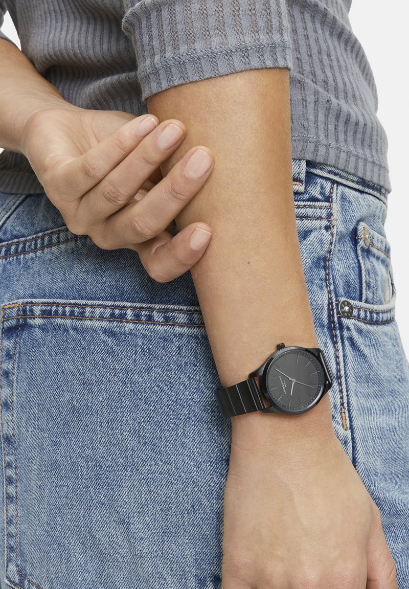 Schwarze analoge Uhr an einem Handgelenk, mit einem runden Ziffernblatt und einfachen Markierungen, kombiniert mit einem schwarzen Metallarmband. Denimstoff im Hintergrund.