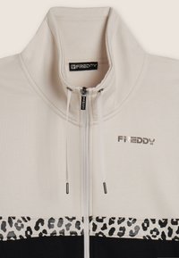 Giacca beige e nera con zip, colletto alto, cordini bianchi, striscia in stampa leopardata e logo metallico "FREDDY" sul petto.