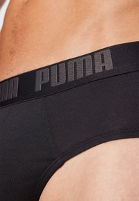 Schwarze Baumwollslips mit elastischem Bund, auf dem der Markenname "PUMA" in Grau zu sehen ist. Nahaufnahme, die die Textur und die Nähdetails hervorhebt.