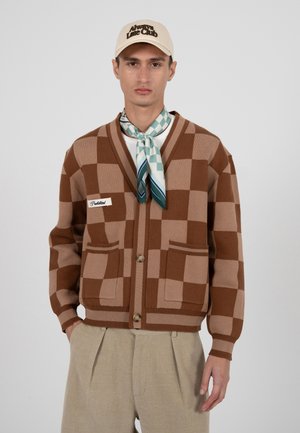 Uomo con cappellino beige, sciarpa a quadri turchese, cardigan marrone a quadri con tasche e pantaloni beige plissettati su sfondo semplice.