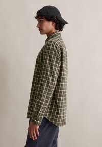 Jongere in een donkergroene geruite shirt, marineblauwe corduroybroek en een zwarte honkbalpet achterstevoren, staande in profiel tegen een neutrale achtergrond.