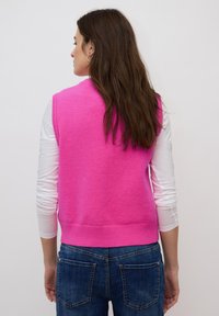 Femme aux longs cheveux bruns portant un pull sans manches rose vif par-dessus un haut à manches longues blanc et un jean bleu, de dos.
