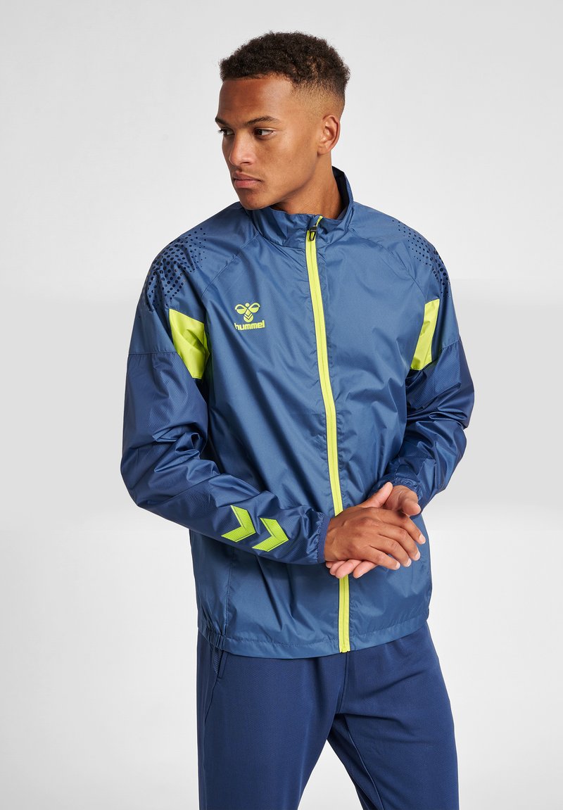 Marinblå lättviktsjacka med neongröna detaljer, dragkedjestängning, detaljerat axelmönster och chevronlogotyp på ärmen.