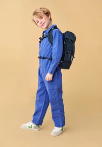 Tutina in denim blu con maniche lunghe, tasche anteriori e cintura regolabile. Zaino nero con motivo a stelle. Scarpe bianche con dettagli verdi.