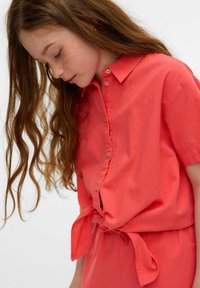 Camicia corallo con maniche corte e vita annodata, caratterizzata da una texture in tessuto morbida e dettagli dei bottoni discreti.