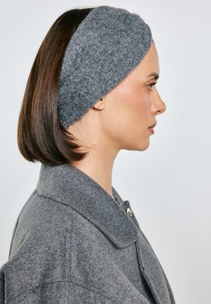 Kobieta w profilu, ubrana w szarą wełnianą opaskę na głowę i pasujący płaszcz, z brązowymi, sięgającymi do ramion, prostymi włosami na jednolitym tle.