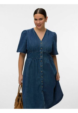 Kvinde iført en blå denimkjole med korte ærmer og knapper, der holder en tanfarvet læderhåndtaske mod en ensartet lys baggrund.