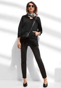 Zwarte tuniek met een keyhole-halslijn, gecombineerd met zwart-witte polkadotbroek. Accessoires zijn een patroon sjaal en een zwarte crossbody tas.
