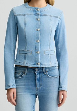 Lyseblå denimjakke med perleknapper, kombineret med matchende lyseblå denimjeans, båret af en stående person.