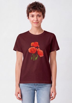Jeune personne aux cheveux courts portant un t-shirt rouge foncé avec trois fleurs de coquelicot orange et un jean bleu clair, debout devant un fond uni.