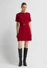 Rood short-sleeve jurk met een gestructureerde silhouet, voorzien van een tailleband en accenten van goudkleurig metaal. Draagt men met zwarte kniehoge laarzen.