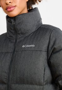 Crna puffer jakna sa ribljim kostima, visokim ovratnikom, zatvaranjem na rajsfšlus i Columbia logotipom na prsima. Tekstura tkanine s kastenim dizajnom.