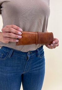 Brūna ādas maks ar gludu tekstūru, rullēta dizaina, šūtām malām un ietītu siksniņu. Turēts vienā rokā uz neitrāla fona.