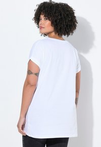 Bílá bavlněná tričko s krátkými rukávy, volným střihem a zaobleným výstřihem, s delším zadním lemem a viditelným tetováním na paži.