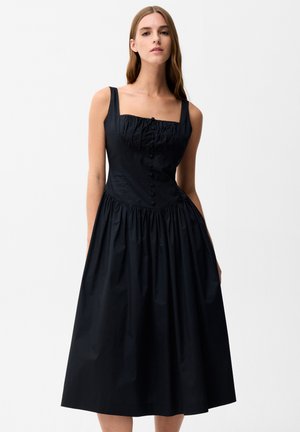 Vrouw in een mouwloze zwarte midi-jurk met geplooide taille, knopen aan de voorkant, aangesloten taille en uitlopende rok tegen een effen witte achtergrond.