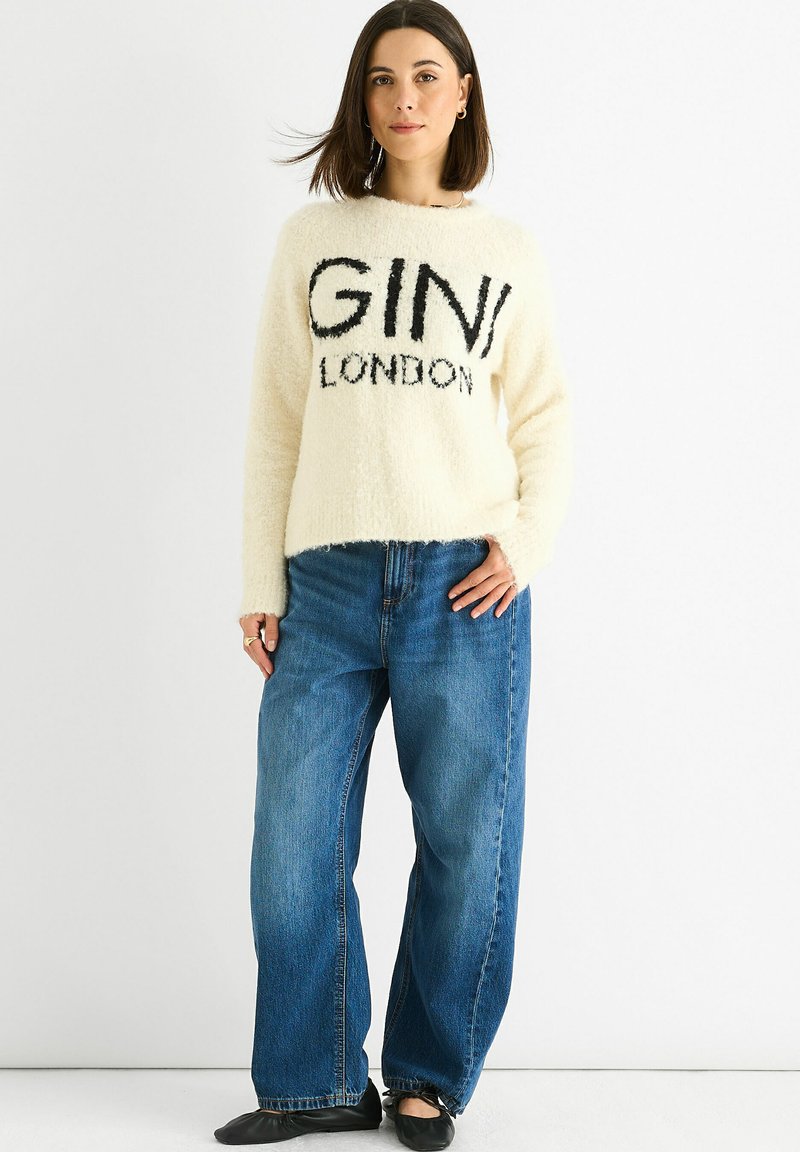Woman wearing a cream fuzzy sweater with "GINI LONDON" text, loose blue jeans, and black shoes, standing against a white background.