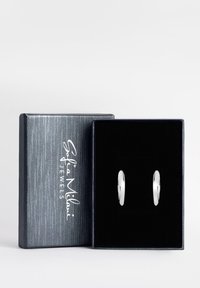 Silverringar i form av hoops visade i en svart sammetsklädd låda med en texturerad mörkblå utsida och vit logotyp på locket.